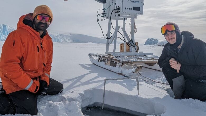 Two men kneel on thick sea ice with measuring equipment on the ice behind them.