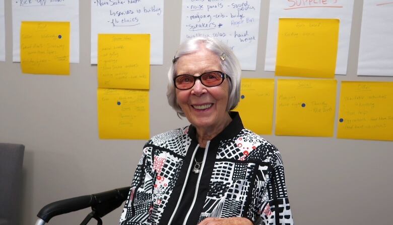 An elderly woman with white hair and glasses smiles at the camera