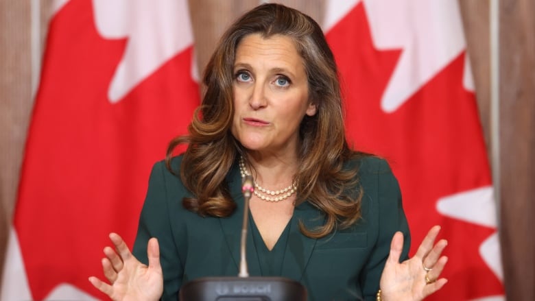 A woman with long brown hair, wearing a green outfit, gestures as she sits at a microphone in front of a Canadian flag.