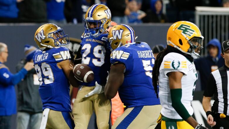 Players in blue jerseys gather on a field while a player in a white jersey walks next to them.