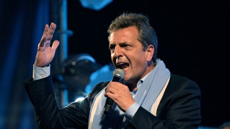 A man in a dark suit holds a microphone while he speaks during a political rally.