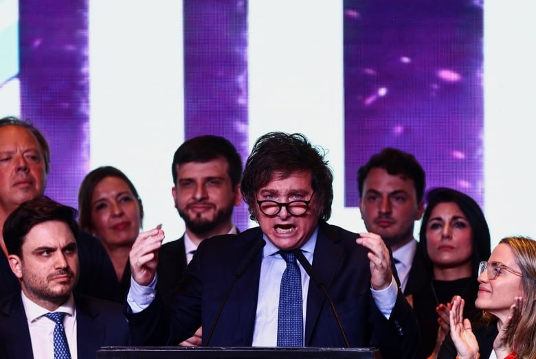 A man stands on a stage flanked by supporters while he addresses a crowd at a political rally.