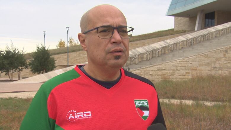 A man wears a colourful shirt with the Palestine flag on it. He is bald and has glasses on as he speaks into a microphone during an interview.