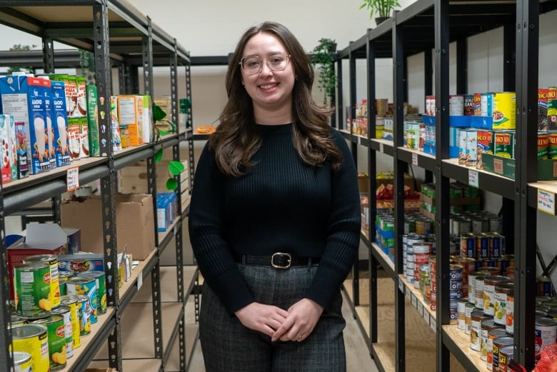 A young woman stands in a room, smiling. Beside her are shelves filled with food.