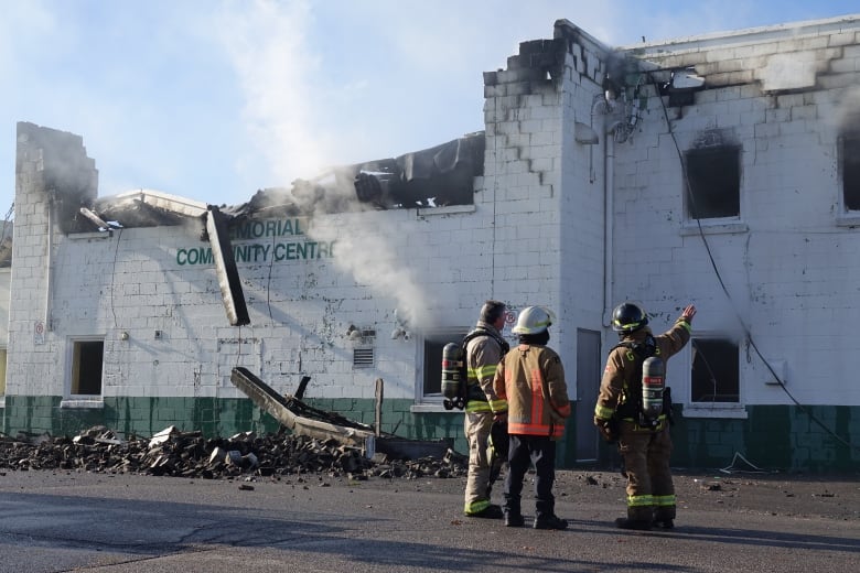 Three firefighters in full gear stand in front of a white and green brick building that's partially collapsed. Smoke is in the air and rubble is one the ground.