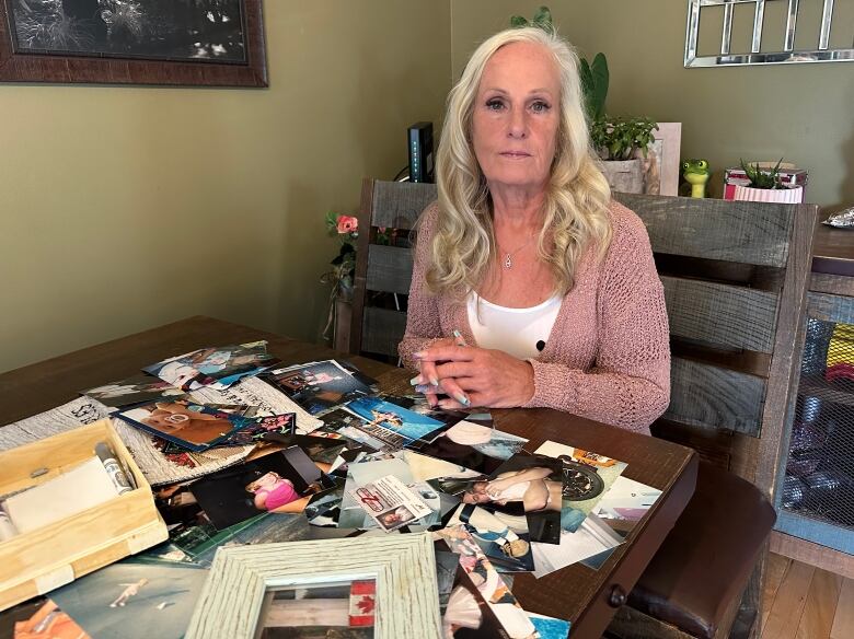 An older woman with blond hair sits at a table covered in photos, looking at the camera.