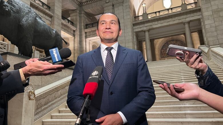 A man stands on the steps inside the Manitoba Legislative Building as microphones are pointed at him.