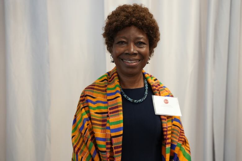 A woman wearing a colourful sweater stands in front of a white curtain.