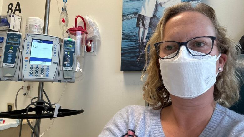 Natalie Kwadrans is wearing a white mask and glasses as she gets her treatment at the Tom Baker Cancer Centre. There is an IV pump beside her.