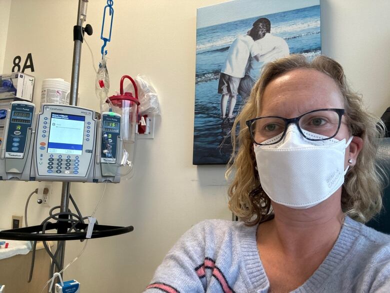 Natalie Kwadrans is wearing a white mask and glasses as she gets her treatment at the Tom Baker Cancer Centre. There is an IV pump beside her.