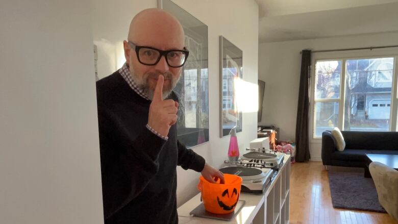 A man with glasses holds a finger to his mouth in a shushing motion while stealing from a Halloween candy bucket with the other hand.