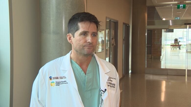 A man in a white doctors coat and scrubs standing in the hallway of a hospital.