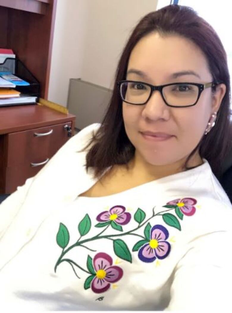 A Cree woman wearing glasses wears a white sweater with a design of Indigenous flowers smiles at the camera in her office.