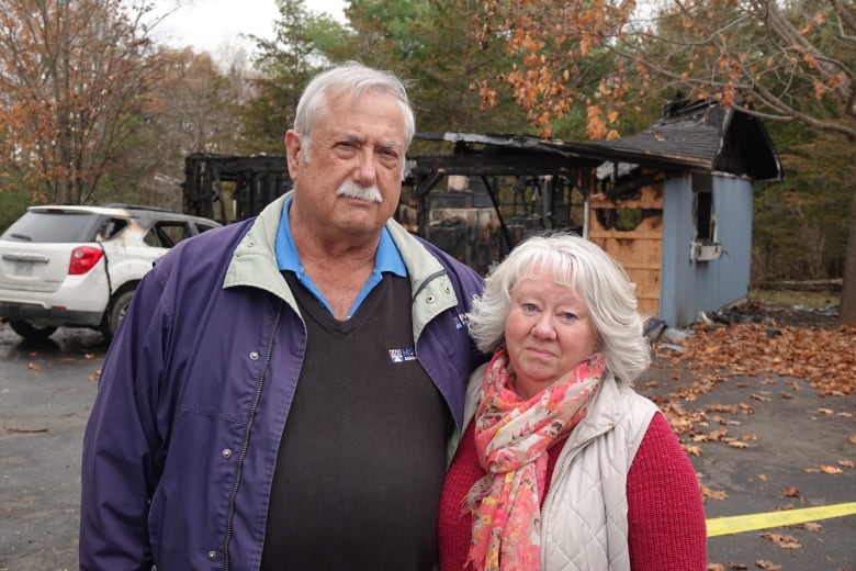 A tall man with white hair and a moustache stands next to a small woman in a white vest and orange sweater. Behind them is a burned down garage and a vehicle with scorch marks on it.