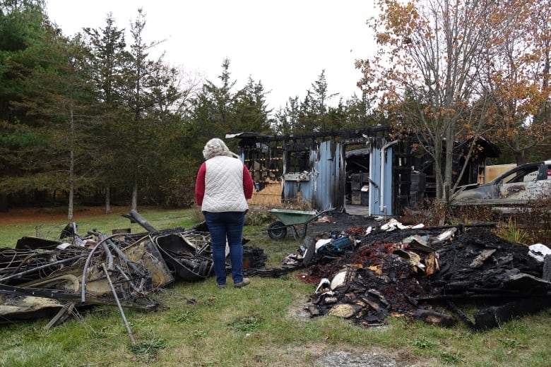 A woman with white hair and wearing jeans and a white puffer vest stands between two blackened piles of fire damaged items. The remains of a bicycle and other twisted bits of metal can be seen among them.