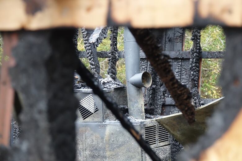 Metal tubes can be seen through a hole in a piece of plywood, which is burned around its edges. The furnace is surrounded by charred wood and ashes.