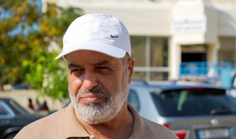 A man in a beige golf shirt and white baseball cap looks off camera, frowning. 