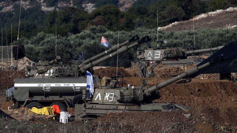 Three tanks, their guns angled up, sit amid some mounds of earth and scrub vegetation.