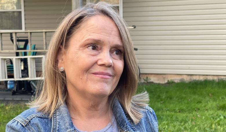 A white woman with shoulder-length dark blonde hair wears a jean jacket and teeshirt. She stands outside in a yard, with a home behind her