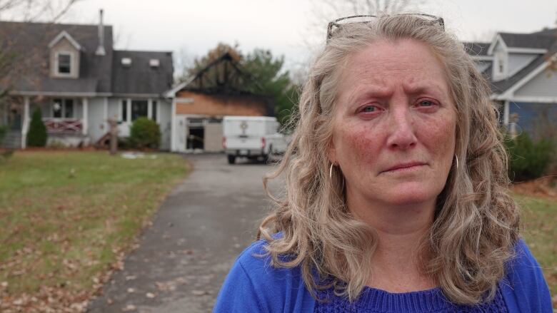 A woman with curly grey-blonde hair, her eyes red from crying, stares at the camera with a sad look on her face. Behind her is the burned remnants of a garage.