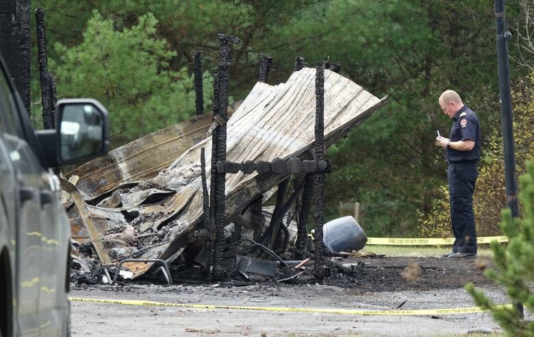 A man in a firefighter's uniform writes on a yellow notepad. He's standing next to the burned wreckage of a wooden wall.