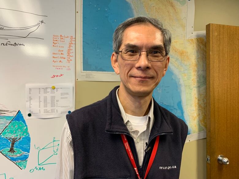 A man wearing glasses and a blue vest stands in front of a map.