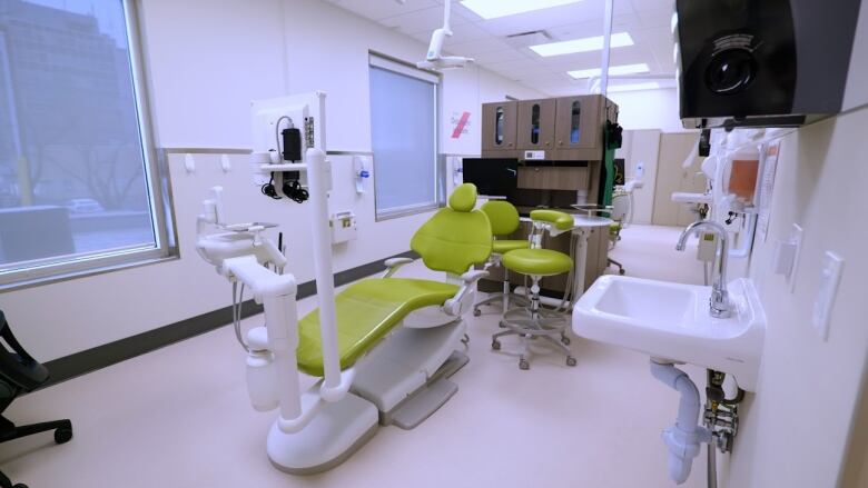 A dental treatment room. White room. Dental workstation in the middle, yellow-greenish in color. A cupboard in the background with some chairs. 