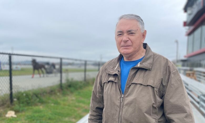 An older man with short hair looks off camera. A horse being raced by a harness driver can be seen out of focus in the background.