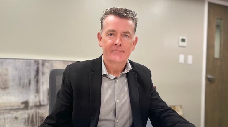 A man with short hair wearing a dress shirt and suit jacket sits at a desk