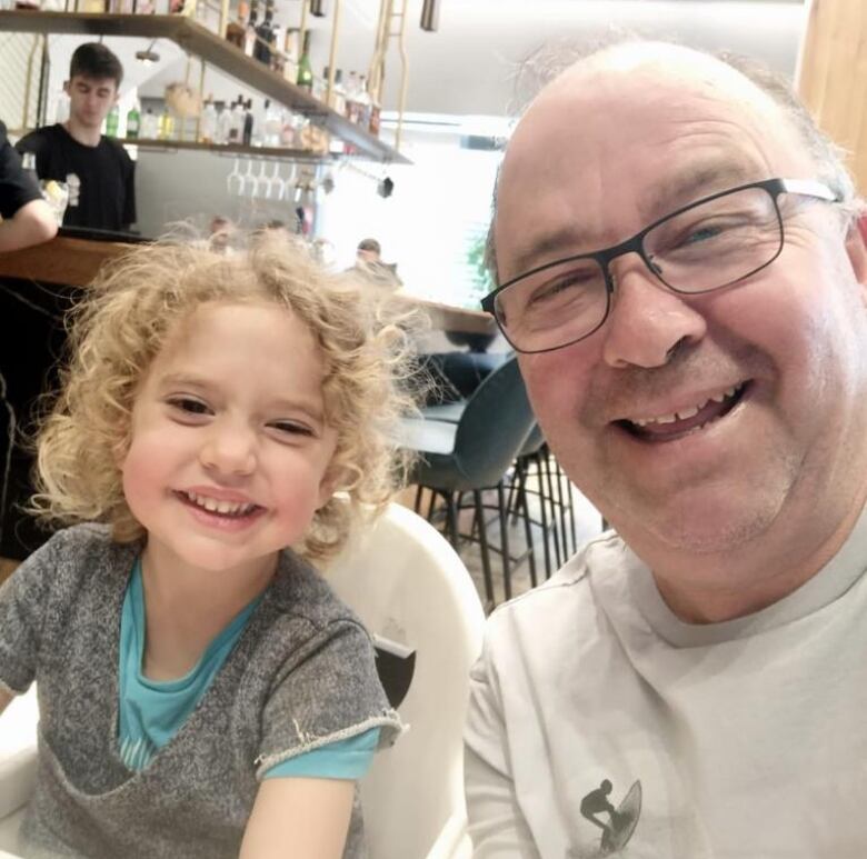 Restaurant selfie with a grinning bald man with glasses and a little girl with curly locks sitting in a high chair.  