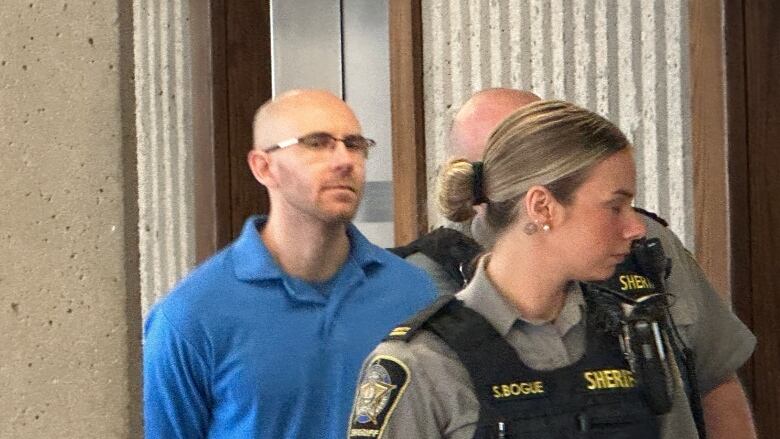 A man wearing a blue collared shirt and glasses is escorted by two police officers. 