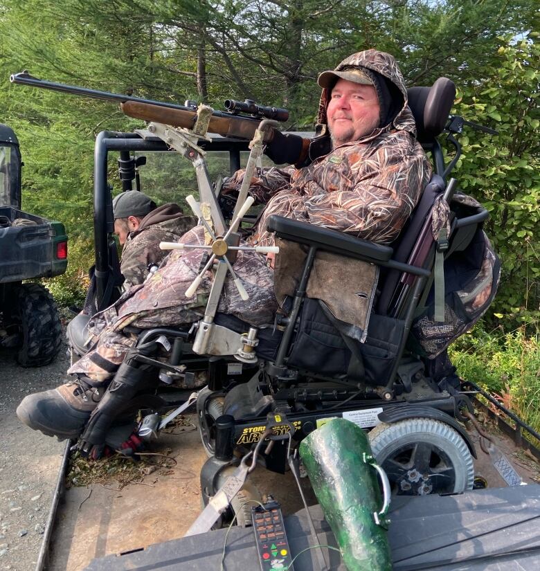 A man wearing camouflage and holding a rifle sits in a wheelchair. The rifle is attached to a stand that mounts it.