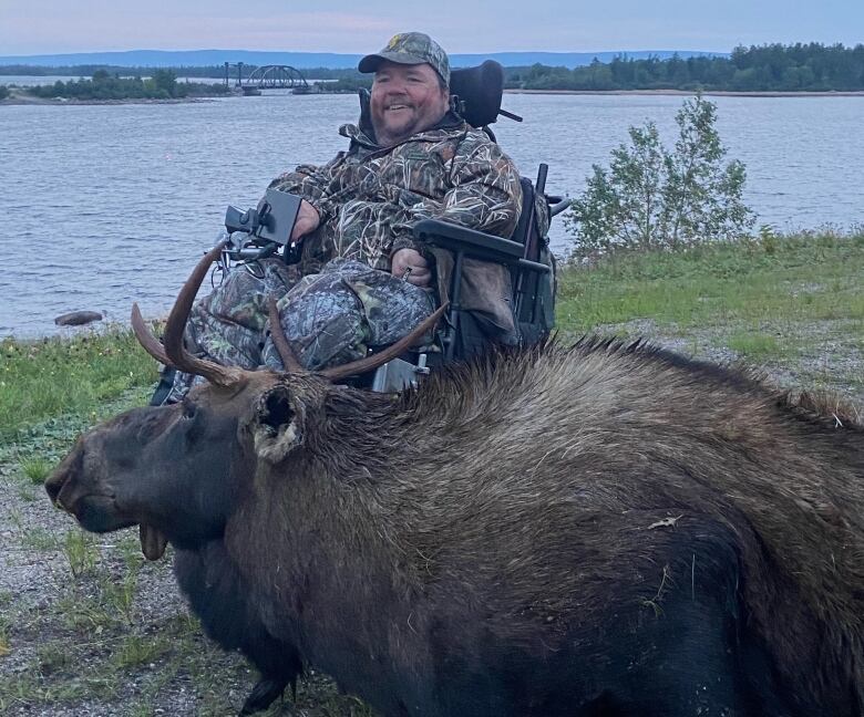 A man sitting in a wheelchair smiles as he sits over a large bull moose.