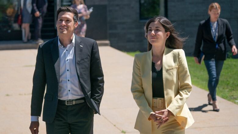 A young man and young woman dressed in business attire stride away from a building.