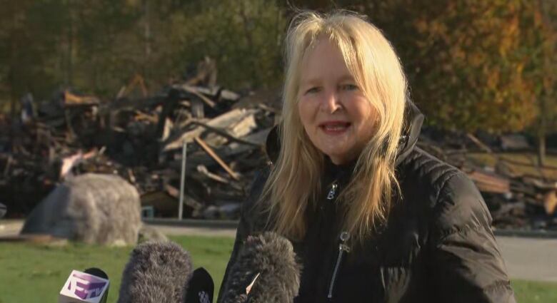 A woman speaks into an array of microphones with fire scorched debris behind her.