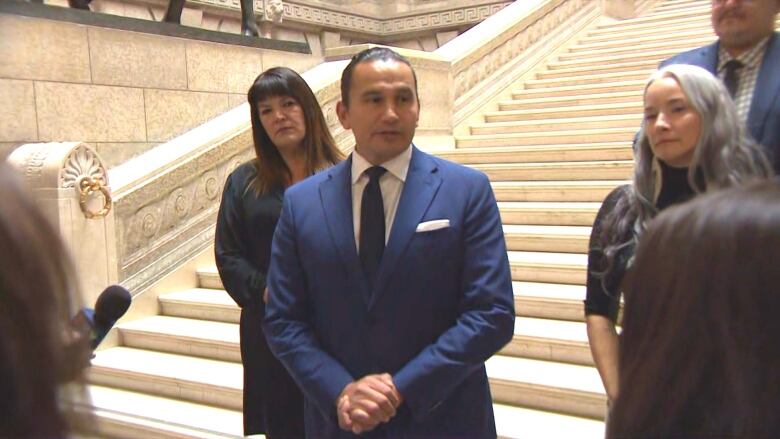 Man with hands clasped together stands in front of the staircase in the rotunda of the Manitoba Legislature.