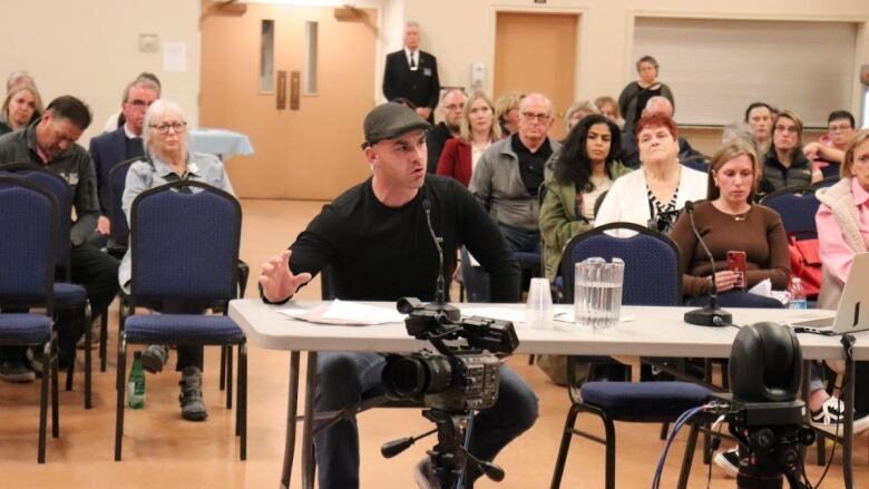 A man in a black shirt, jeans and a hat sitting at a table with a microphone in front of him with people sitting behind him.