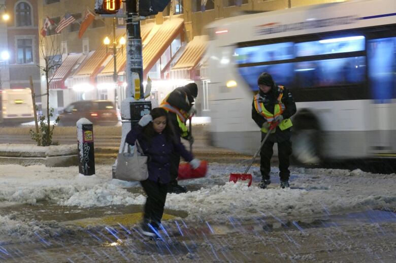 A woman crosses a snowy street and crews in bright vests shovel a snowy sidewalk in the background.