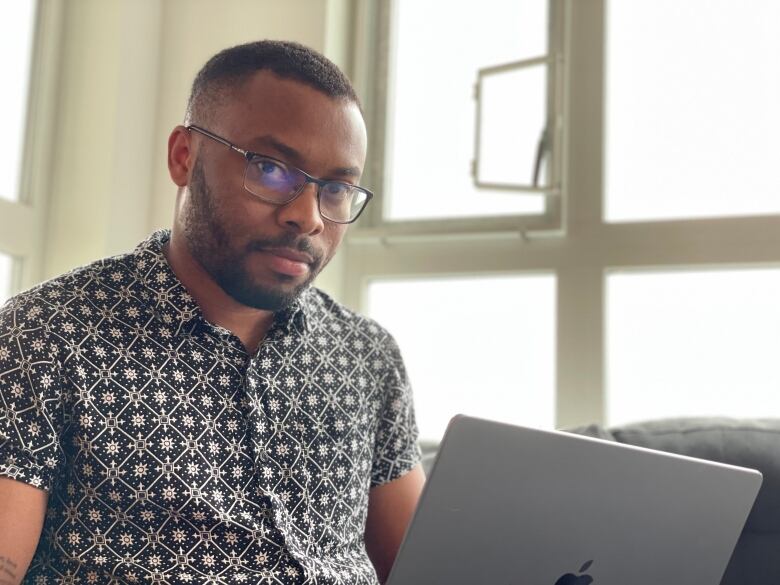 A man seated in front of a window holds a laptop on his lap. 