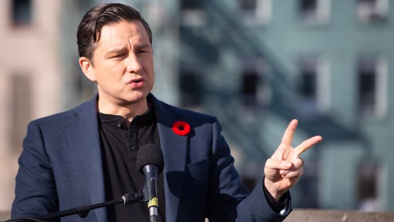 Pierre Poilievre, wearing a suit jacket over a t-shirt, speaks into a microphone. 