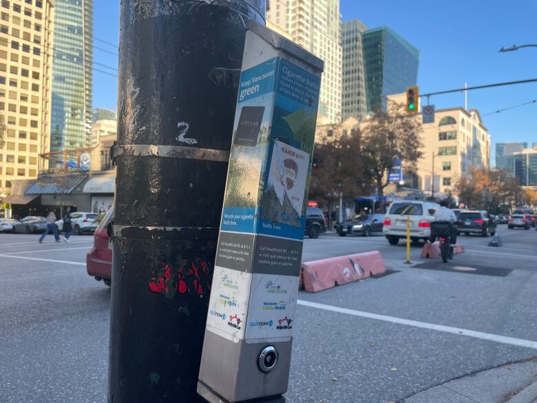 A slender steel box covered in stickers attached to a light standard on the corner of a busy downtown Vancouver interseciton.