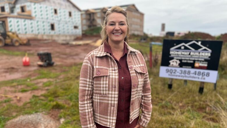 Smiling woman in a brown and white plaid jacket stands in front of an apartment building under construction. 