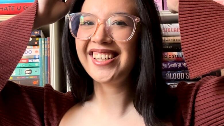 Young Filipino-Irish woman with long black hair and a red top on holding a stack on books on her head in front of bookshelves.