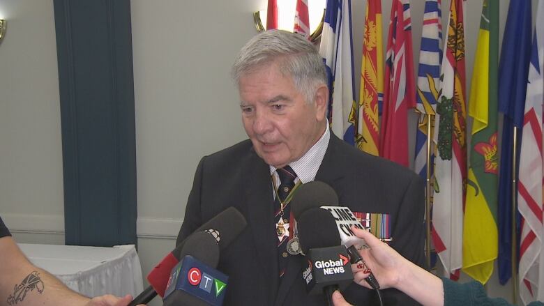 An older man with light skin and grey hair is wearing a three-piece suit, with a poppy on his lapel and military medals over his heart. Provincial flags are behind him.