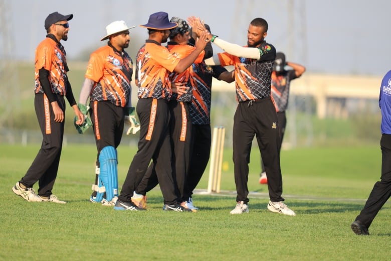 A team of cricket players wearing orange shirts huddle in a group and high five each other on a cricket pitch.