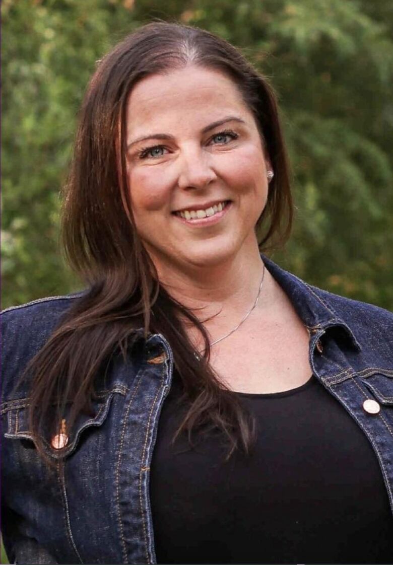 Woman smiles at camera. Stands in front of trees.