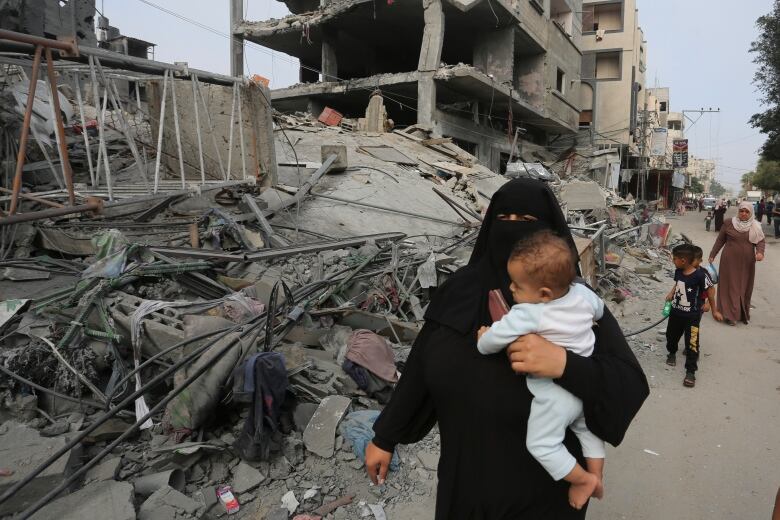 A woman in a black garment covering her head and face carries a baby as she walks past a large pile of concrete rubble and destroyed buildings.