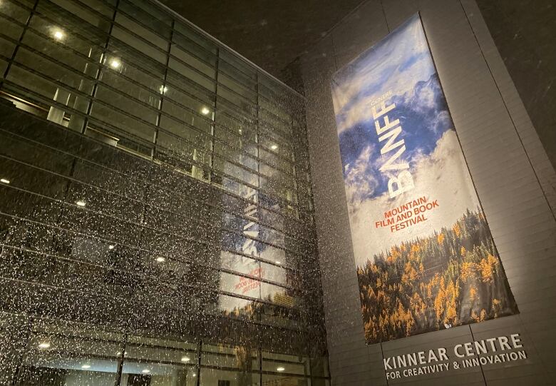 Snow falls at night in front of a large building with a big book festival banner hanging down its front.