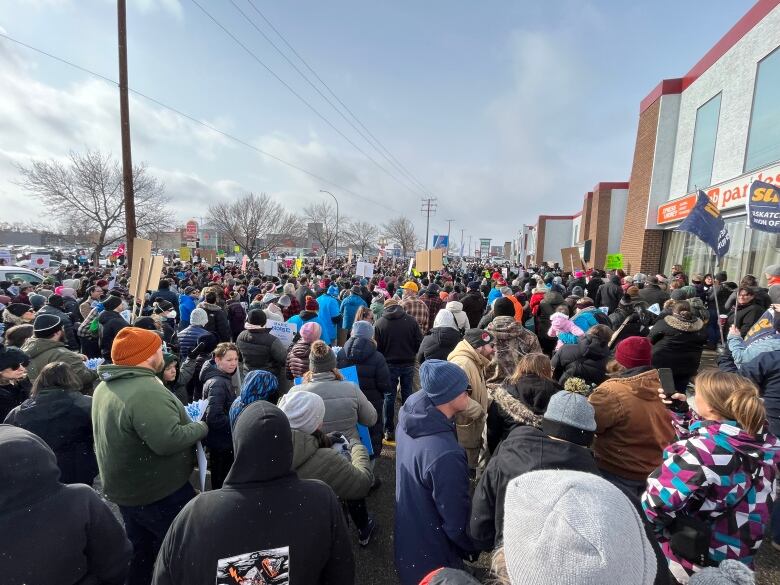 Hundreds of people are packed wearing winter clothing in a crowd outside a strip mall