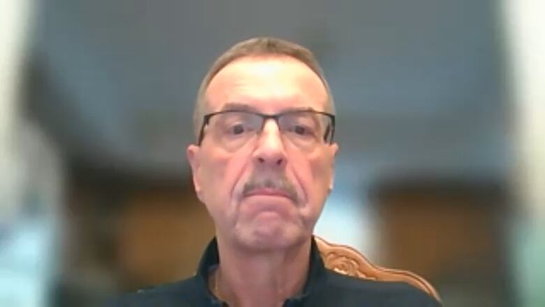 A man with glasses and a moustache looks directly at the camera. The background is blurred.